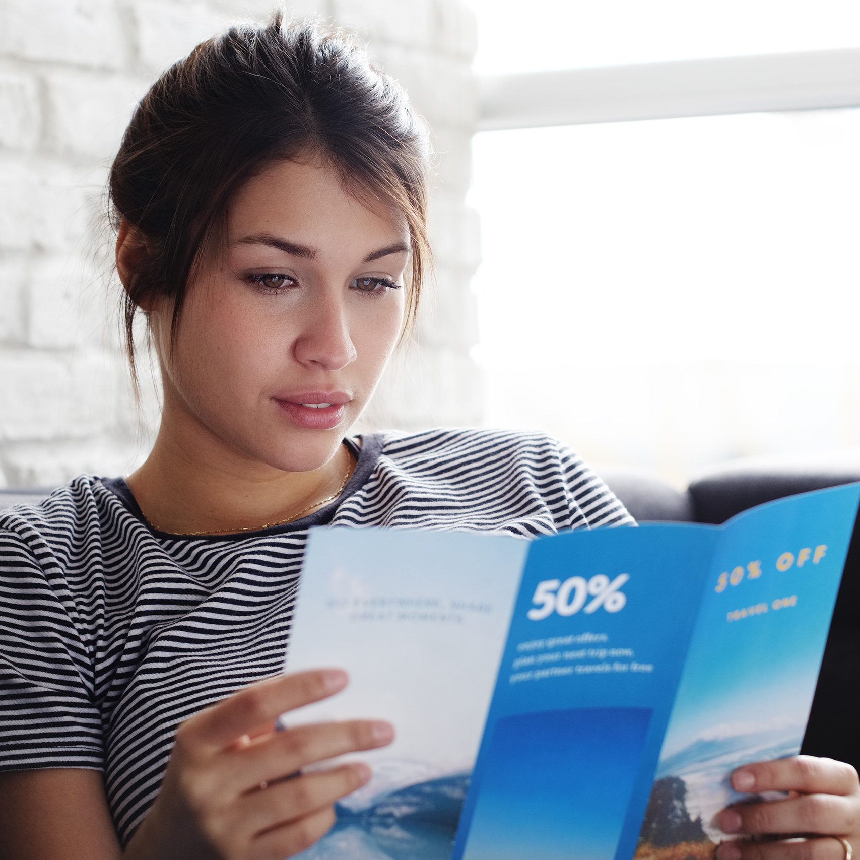 Young hispanic woman laying on couch at home, holding a travel flyer and planning next trip. Copy space on window.